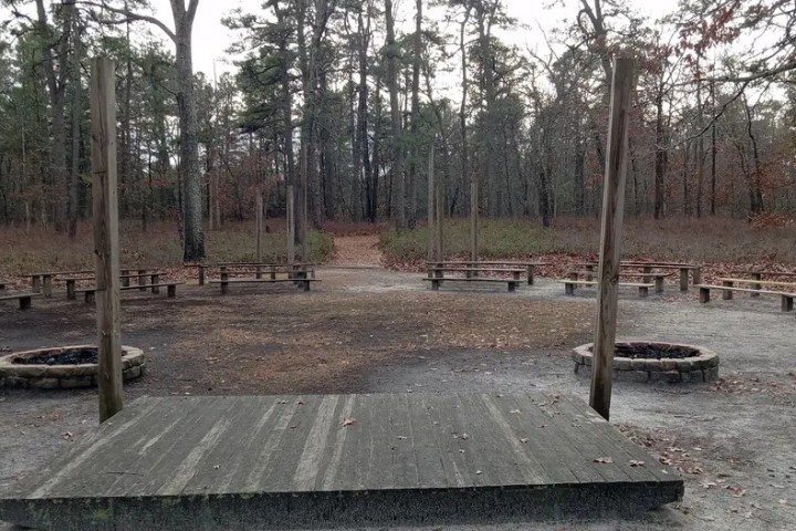 Outdoor amphitheater in a forest with stage, benches, and fire pits.