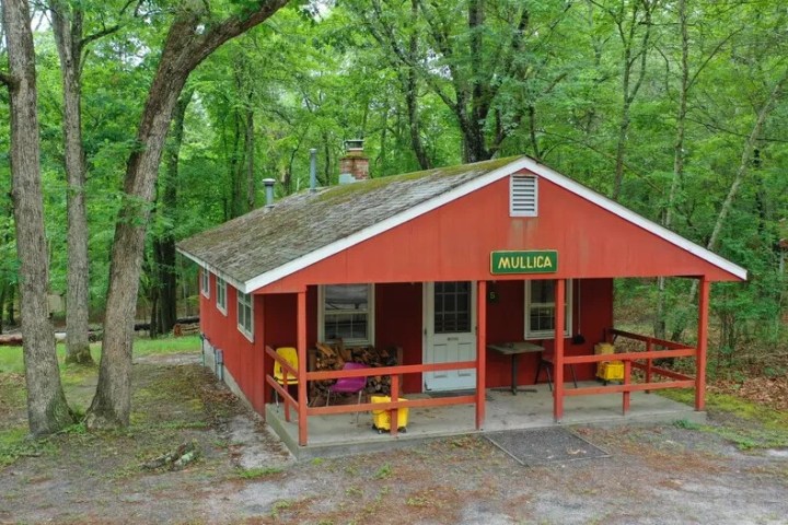 Red cabin in a forest with a porch and sign reading 'Mullica'.