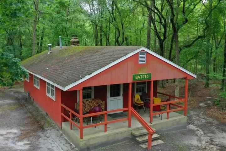 Red cabin in the forest with front porch, sign labeled 'BATSTO', and chairs in front.