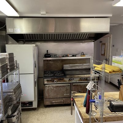 Commercial kitchen with stove, oven, fridge, and shelves with cooking supplies and utensils.