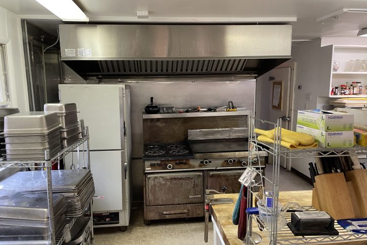 Commercial kitchen with stove, oven, fridge, and shelves with cooking supplies and utensils.