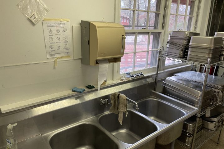 Commercial kitchen sink with paper towel dispenser, cleaning supplies, and stacked trays by the window.