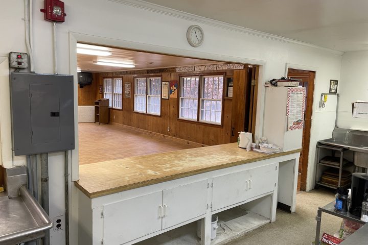 Empty kitchen with a wooden counter and view into a wooden-floored room with windows.