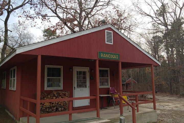 Red cabin with 'Rancocas' sign, chairs stacked outside, surrounded by leafless trees.