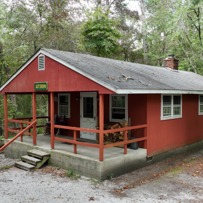 Red cabin with porch in forest setting, surrounded by trees.