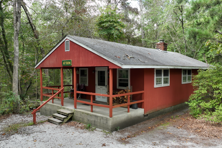 Red cabin with porch in forest setting, surrounded by trees.