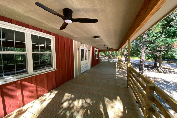 Covered wooden porch with ceiling fan, red siding, and railing overlooking wooded area.