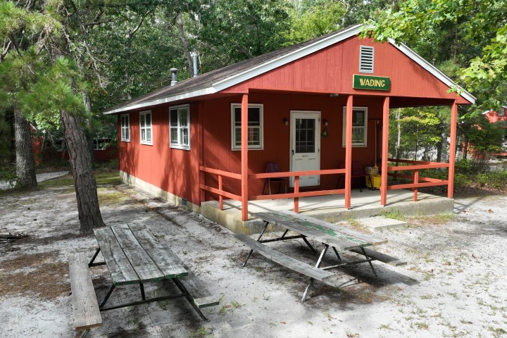 Red cabin with porch and 'WADING' sign, surrounded by trees, with picnic tables in front.